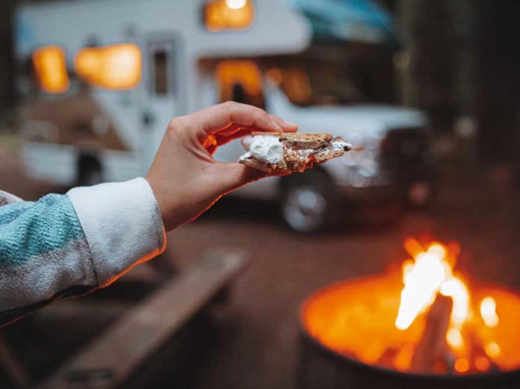 Hand holding a smores treat in front of a fire in a fire pit and RV