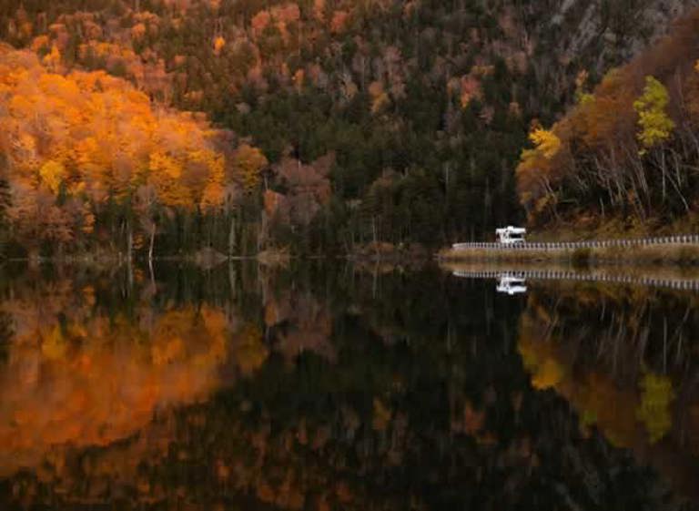 RV driving down road next to a lake and a forest in autum.
